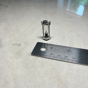 May include: A small silver hourglass with a black top and a clear glass bulb. The hourglass is approximately 2.5 cm tall and is sitting on a white surface next to a ruler.