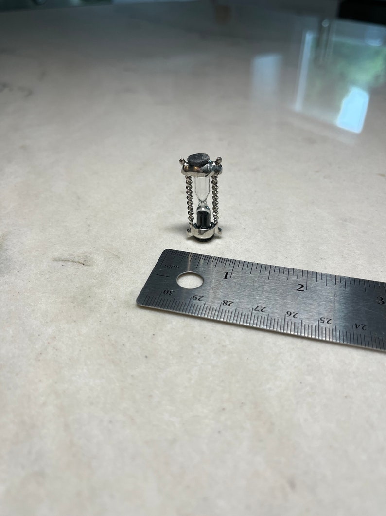 May include: A small silver hourglass with a black top and a clear glass bulb. The hourglass is approximately 2.5 cm tall and is sitting on a white surface next to a ruler.