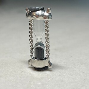 May include: A small silver hourglass with a chain detail. The hourglass is filled with black sand.