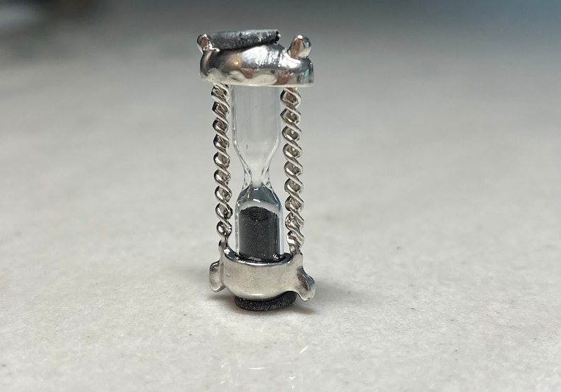 May include: A small silver hourglass with a chain detail. The hourglass is filled with black sand.