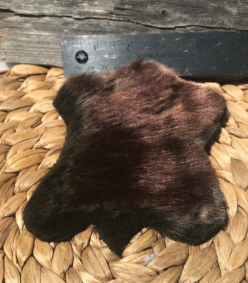K&ouml;nnte beinhalten: Ein dunkelbraunes Kunstfellst&uuml;ck mit leicht unregelm&auml;&szlig;iger Form. Das Fell ist weich und flauschig mit einem leichten Glanz. Das St&uuml;ck ist etwa 10 cm breit und 12,5 cm lang.
