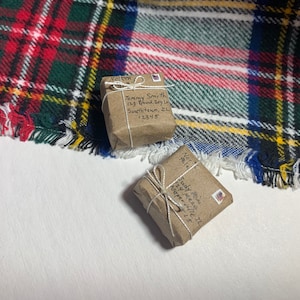 May include: Two small, brown packages wrapped in twine and tied with a bow. The packages are sitting on a red, green, and white plaid fabric. The packages have handwritten addresses on them.