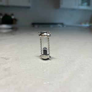 May include: A small silver hourglass with a chain detail. The hourglass is filled with sand and is sitting on a white surface.