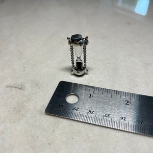 May include: A small silver hourglass with a black top and bottom. The hourglass is approximately 2.5 cm tall and is resting on a ruler.