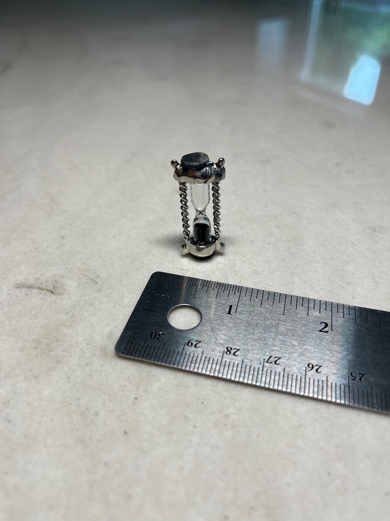 May include: A small silver hourglass with a black top and bottom. The hourglass is approximately 2.5 cm tall and is resting on a ruler.