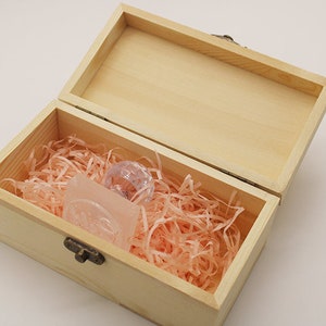 May include: A wooden box with a hinged lid, filled with pink shredded paper and two small, clear glass objects. The box has a brass latch.