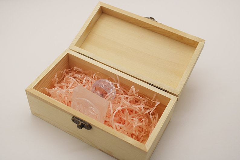 May include: A wooden box with a hinged lid, filled with pink shredded paper and two small, clear glass objects. The box has a brass latch.