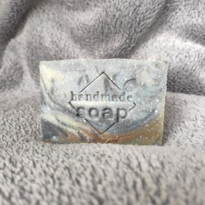 May include: A bar of handmade soap with a gray, white, and brown marbled design. The soap has a diamond-shaped stamp that says "handmade soap".