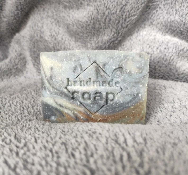 May include: A bar of handmade soap with a gray, white, and brown marbled design. The soap has a diamond-shaped stamp that says "handmade soap".