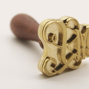May include: A gold-colored metal stamp with a wooden handle. The stamp features a decorative, stylized letter "L" design.
