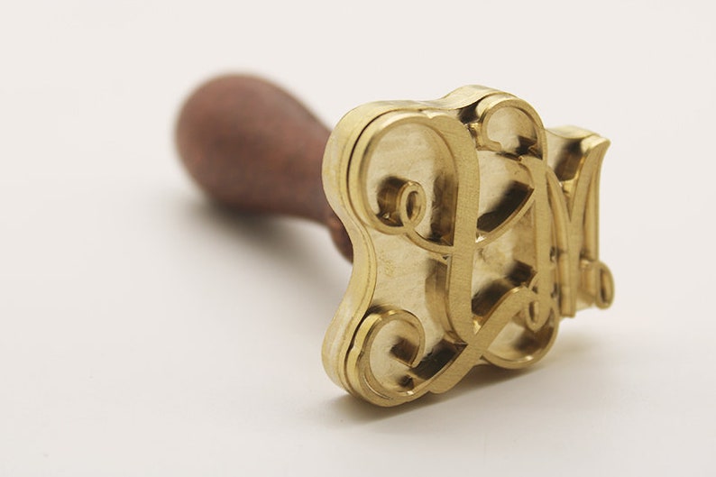May include: A gold-colored metal stamp with a wooden handle. The stamp features a decorative, stylized letter "L" design.
