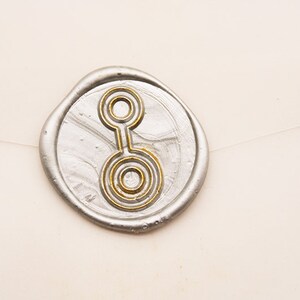 Könnte beinhalten: Ein silberner Wachssiegel mit einem goldfarbenen Design aus konzentrischen Kreisen und Linien. Der Siegel befindet sich auf einer cremefarbenen Oberfläche, vermutlich Briefpapier. Das Design ist zentriert und detailliert.