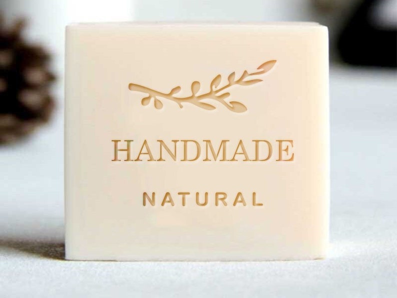 May include: A white bar of soap with a simple twig design and the words "HANDMADE NATURAL" embossed on the front.