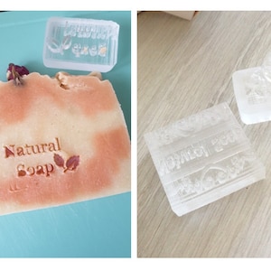 May include: Three clear plastic soap stamps with raised designs. One stamp says "Natural Soap" with two leaves. Another stamp has a floral design. The third stamp has a design with a heart and a banner.