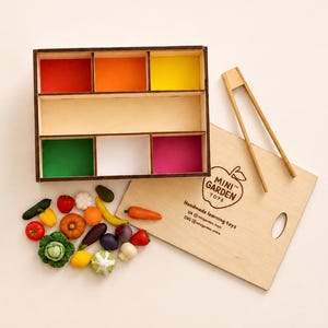 Peut inclure: Un plateau de tri en bois avec six compartiments colorés, un ensemble de légumes miniatures et des pinces en bois. Le couvercle du plateau porte le texte MINI GARDEN TOYS, Handmade learning toys et des identifiants imprimés en noir.