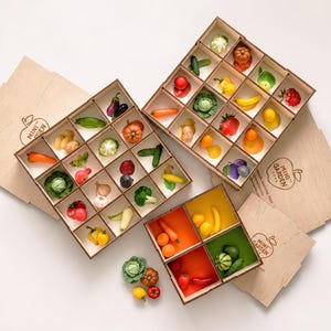 Peut inclure: Trois boîtes en bois avec des séparateurs contenant des figurines miniatures de fruits et légumes. Les boîtes portent le texte MINI GARDEN TOTS et une icône de pomme. La collection comprend divers produits colorés comme des carottes, des tomates et des légumes verts, avec quelques pièces éparpillées à l'extérieur.