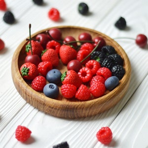 May include: A wooden bowl filled with a variety of fresh berries, including strawberries, raspberries, blueberries, and cherries. The bowl is on a white wooden surface.