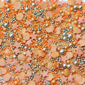 Könnte beinhalten: Eine Mischung aus orangefarbenen, goldenen und silbernen Streuseln. Die Streusel haben die Form von Sternen, Kürbissen und kleinen Stäbchen.