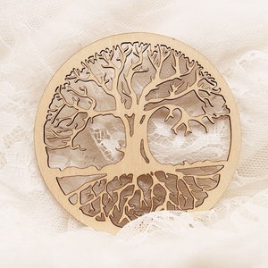 May include: A circular wooden tree of life design. The tree is intricately cut out, revealing a white, lacy background. The wood has a light, natural finish. The design is circular and could be a coaster or decorative piece.
