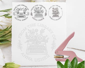 May include: A dusty rose metal embosser tool positioned next to a white textured paper sheet. The paper displays three black circular ink stamp designs featuring a typewriter with floral arrangements and the text From The Library Of.