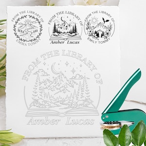 Könnte beinhalten: Ein grünes Prägewerkzeug aus Metall neben einem weißen Blatt Papier mit drei kreisförmigen schwarzen Stempeln und einem großen Prägemotiv. Die Designs zeigen Berge, Wälder und Bücher mit dem Text FROM THE LIBRARY OF.