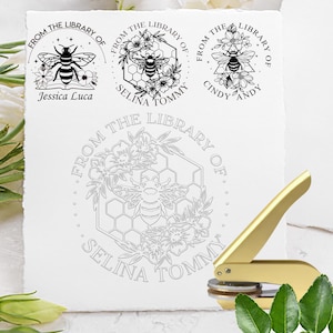 May include: A gold metal embosser tool positioned next to a white paper sheet featuring three black ink stamp impressions of bees, flowers, and honeycombs with the text From The Library Of Jessica Luca, Selina Tommy, and Cindy Andy.
