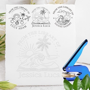 Puede incluir: Una herramienta de estampado de metal azul brillante colocada junto a una hoja de papel blanco con cuatro diseños de sello de tinta negra circulares. Los diseños incluyen ilustraciones de palmeras, olas, soles y el texto From The Library Of.