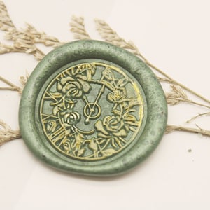 Può includere: Un sigillo di cera verde con un designo floreale dorato e un quadrante dell'orologio al centro.