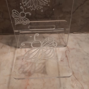 May include: A clear acrylic phone stand with a laser-etched design of a sunflower and two butterflies. The stand is designed to hold a phone upright.