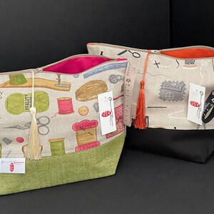 May include: Two zippered pouches with a sewing theme. One pouch is beige with a green bottom and features a sewing machine, scissors, spools of thread, buttons, and a tassel. The other pouch is beige with a black bottom and features a sewing needle, pins, and a measuring tape.