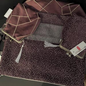 May include: A set of three mauve-coloured accessories including two small pouches with metal frames and tassels, and one large rectangular clutch with a textured leaf-like pattern. A white tag with red text and a logo is attached to one pouch.