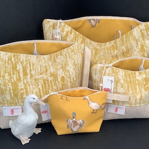 May include: Four yellow fabric zippered pouches with a white duck print. The pouches are lined with a solid yellow fabric. The largest pouch has a white fabric strap. A white porcelain duck figurine is in the foreground.