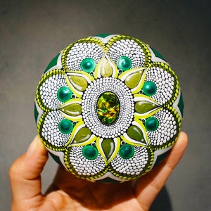 Puede incluir: Una piedra decorativa pintada a mano con un diseño de mandala. La piedra es principalmente verde con acentos blancos, amarillos y verde esmeralda. El centro tiene una gran gema ovalada iridiscente. El diseño se compone de puntos y patrones florales.