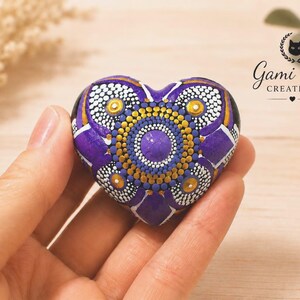 May include: A heart-shaped stone painted with a purple, gold, and white mandala dot art pattern. The design features a central purple dot surrounded by concentric circles of gold and white dots, with purple petal shapes radiating outward.
