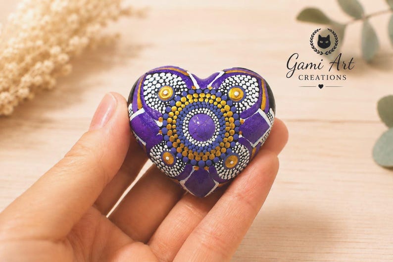 May include: A heart-shaped stone painted with a purple, gold, and white mandala dot art pattern. The design features a central purple dot surrounded by concentric circles of gold and white dots, with purple petal shapes radiating outward.