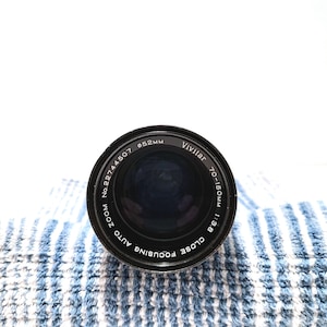 May include: A black Vivitar 70-150mm f/3.8 auto zoom focusing camera lens. The lens has a 52mm diameter and is set against a blue and white striped background. The lens is a close-up lens.