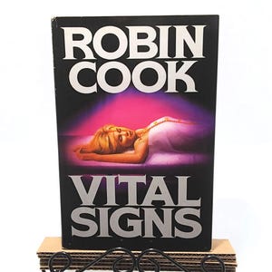 May include: A hardcover book titled "VITAL SIGNS" by Robin Cook. The cover features a woman lying down with a purple and pink background. The book is displayed on a decorative black metal and cardboard stand.
