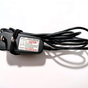 May include: A black Samsung travel adapter with a black cord, coiled and secured with a black strap. The adapter has a white label with the word "CAUTION" in red. The text "DRY LOCATION USE ONLY" is also visible.