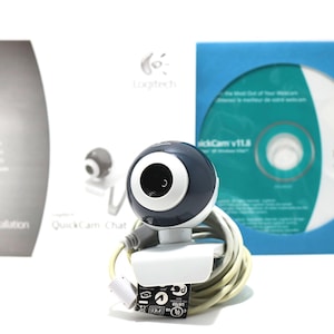 May include: A Logitech QuickCam Chat webcam with a dark blue and white design, coiled cable, and a white base. The image includes a CD-ROM, a manual, and a smaller webcam model. The CD-ROM is in a blue sleeve.