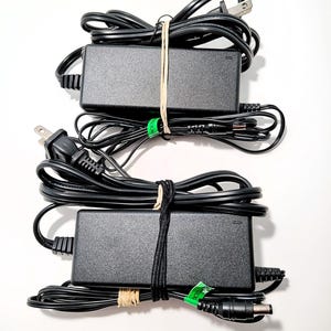 May include: Two black power adapters with attached cords and plugs. Each adapter is rectangular and has a black cord wrapped around it, secured with a rubber band. One adapter has a two-prong plug, and the other has a three-prong plug.