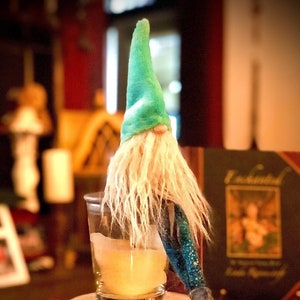 May include: A whimsical gnome figurine with a teal hat and long white beard. The gnome is perched near a glass with a yellow liquid, resting on a white stone base surrounded by dried botanicals. The gnome's outfit includes a blue and green patterned sleeve and a gray shoe.
