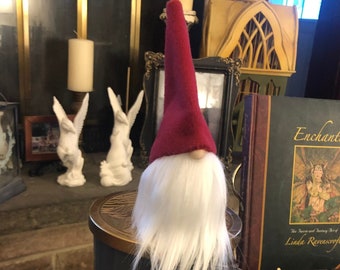 May include: A decorative gnome figure with a red hat and white beard, perched on a candle holder. The gnome is dressed in a plaid jacket and grey felt shoes. The display includes pine cones, dried fruit, and a book titled "Enchanted".
