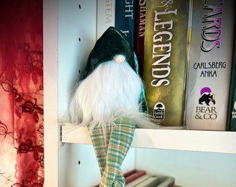 May include: A decorative gnome with a green velvet hat and a long white beard sits on a shelf. The gnome's legs are made of green and tan plaid fabric. The shelf is filled with books, including titles like "LEGENDS" and "MARVEL".