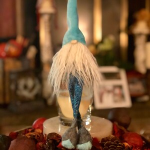 May include: A whimsical gnome figurine with a light blue pointed hat and a long, white beard. The gnome's legs are a dark blue with white dots. The figurine is standing on a decorative tray with potpourri and pinecones.