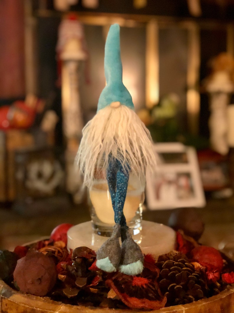 May include: A whimsical gnome figurine with a light blue pointed hat and a long, white beard. The gnome's legs are a dark blue with white dots. The figurine is standing on a decorative tray with potpourri and pinecones.