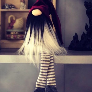 May include: A handmade gnome doll with a burgundy pointed hat, black and white beard, and striped legs. The gnome's feet are dark gray and it is sitting on a shelf. The text "ELDER STYX" is above the gnome.