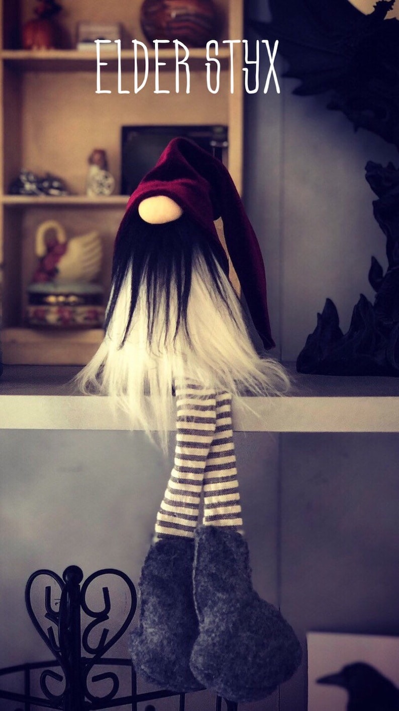 May include: A handmade gnome doll with a burgundy pointed hat, black and white beard, and striped legs. The gnome's feet are dark gray and it is sitting on a shelf. The text "ELDER STYX" is above the gnome.