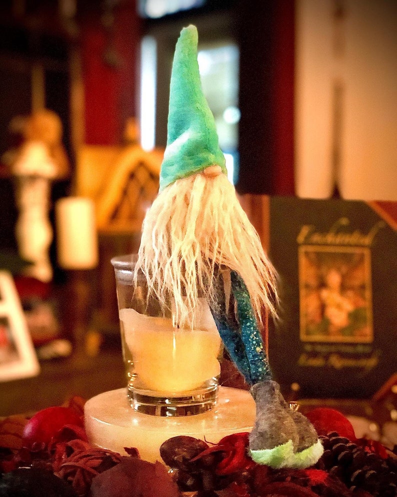 May include: A whimsical gnome figurine with a teal hat and long white beard. The gnome is perched near a glass with a yellow liquid, resting on a white stone base surrounded by dried botanicals. The gnome's outfit includes a blue and green patterned sleeve and a gray shoe.
