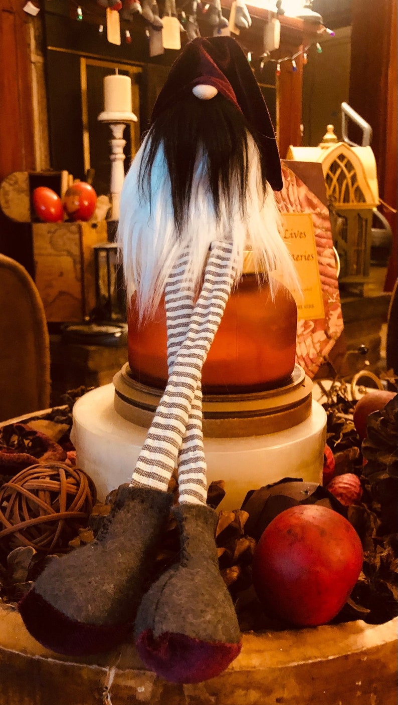 May include: A decorative gnome figurine with a burgundy hat, long black beard, and white fur. The gnome has striped legs and gray boots with purple soles. The figurine is displayed on a white and brown base, surrounded by decorative items.