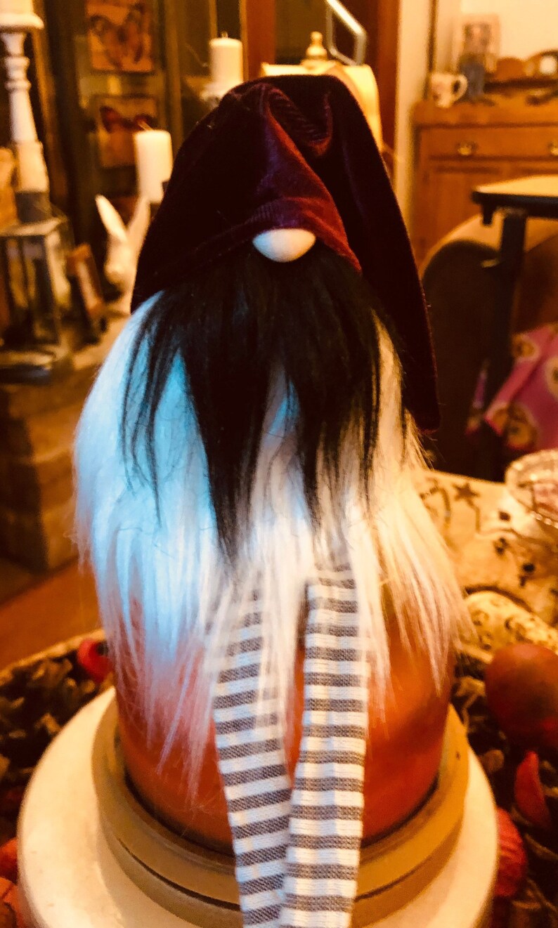 May include: A decorative gnome figurine with a burgundy velvet hat and a long white and black beard. The gnome has a small wooden nose and a striped scarf. The figurine is orange and white.